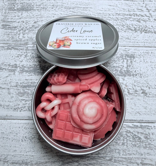 A round metal tin of wax melts shaped like bakery and dessert pieces, with the lid leaning behind it. The label reads Traverse City Wax Co.